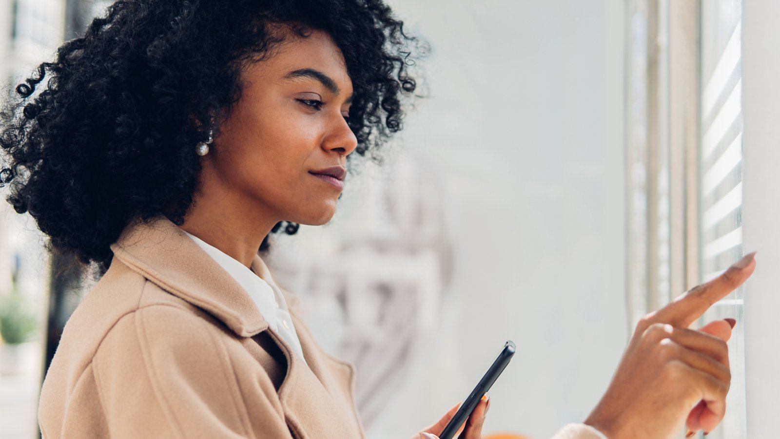 Side view of a confident adult woman using touch screen terminal on street while holding her phone.